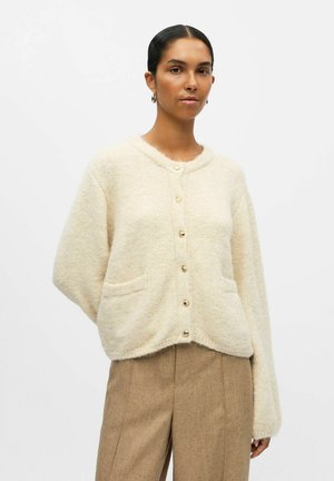 Krämfärgad stickad cardigans med stora knappar, sidofickor, avslappnad passform och textilstruktur. Bärs över beige, strukturerade byxor.