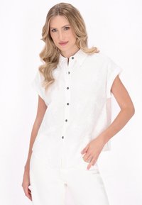 Weiße kurzärmlige Bluse mit durchgeknöpftem Front, die über eine subtile Blumenstruktur und dunkle Knöpfe verfügt, kombiniert mit weißen Hosen.
