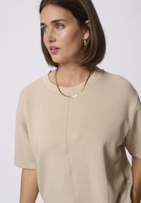 T-shirt oversized beige avec un col rond, des manches courtes et une finition texturée subtile. Accentué par un collier fin en or.