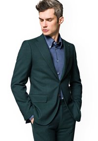 Ciemnozielony garnitur uszyty na miarę z teksturowanej tkaniny, połączony z niebieską koszulą z guzikami. Zauważalne detale to klapa w kształcie litery V oraz kieszenie z przodu.