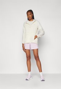 Leichte, cremige Windjacke mit Kapuze, Fronttaschen und Kordelzug in der Taille, kombiniert mit rosa Shorts und sportlichen Schuhen.