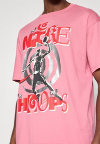 Ροζ βαμβακερή μπλούζα t-shirt με ασπρόμαυρη γραφική παράσταση ενός μπασκετμπολίστ α, με κόκκινο και ασημί κείμενο "NIKE HOOPS," και μικρές αναπαραστάσεις κατσικών.