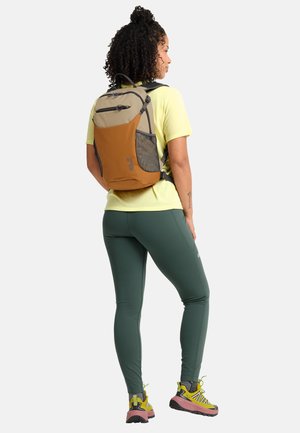 Femme portant des leggings verts, un t-shirt jaune, des baskets jaunes et roses, et un sac à dos bicolore marron, debout devant un fond blanc.