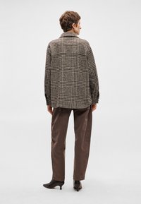 Brūnas pārbaudes raksta pārslēgta jakas, izgatavota no strukturēta auduma, kopā ar brūniem biksēm un melniem augstiem papēžiem, skatīta no aizmugures.