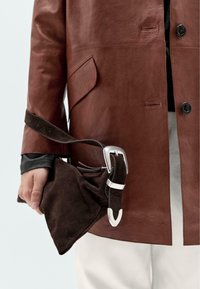 Brown leather jacket with a pointed collar and button details. The hand holds a dark brown suede bag with a silver hardware strap.