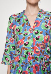 Vestido floral azul com decote em V, mangas 3/4 e saia em camadas. O padrão inclui flores verdes, vermelhas e rosas com folhas castanhas.