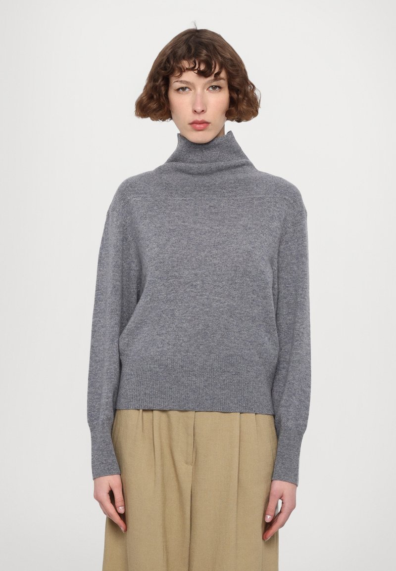Grå genser med høy hals laget av strikket stoff, med ribbestrikket krage og mansjetter, sammen med beige plissert bukser. Enkel og koselig design.