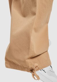 Beige bomullbukser med snøring ved kanten. Glatt tekstur og løs passform, kombinert med hvite sneakers med svarte detaljer.