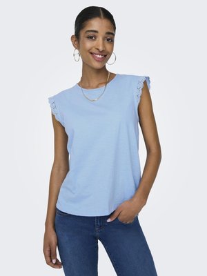 Ung kvinde smilende, iført en lyseblå ærmeløs top med blonder og mørkeblå jeans, hånd i lommen på neutral baggrund.