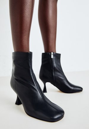 Bottines en cuir noir à bouts pointus et talons blocs courbés portées sur des jambes à la peau foncée, debout sur un sol clair.
