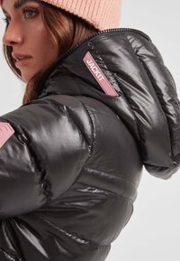 Mujer vestida con una chaqueta acolchada negra brillante con una etiqueta rosa "JACK1T" en la capucha y un gorro de punto rosa, mirando hacia la izquierda.