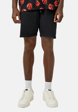 Personne portant un short noir, des chaussettes blanches, des baskets blanches et une chemise noire avec des motifs de roses rouges, debout devant un fond uni.