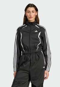 Giacca a vento nera con strisce bianche, chiusura con zip, vita elasticizzata e logo in evidenza. Materiale leggero con una superficie liscia.