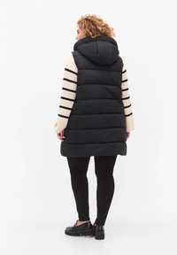 Svart puffer-väst med huva, horisontell quiltad design, kombinerad med en randig långärmad topp och svarta byxor, bärs med plattformsskor.
