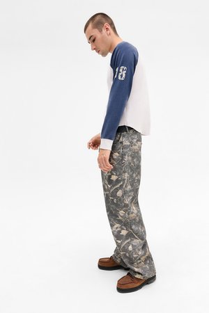 Jeune homme se tenant de profil, portant une chemise à manches longues blanche et bleue, un pantalon ample à motif camouflage et des chaussures marron sur fond blanc.