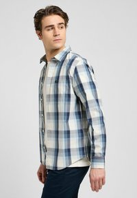 Camicia a quadri blu e bianca con bottoni, realizzata in cotone, presenta una tasca sul petto e maniche lunghe, con orlo curvo e una leggera texture.