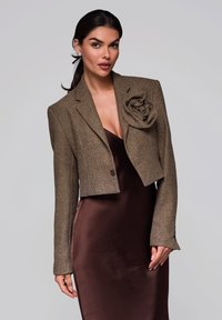 Avskuret brunt tweedjacka med en stor blommig accent, med knappar och långa ärmar, i kombination med en mörk satinklänning.