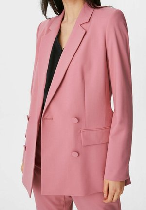 Frau trägt einen maßgeschneiderten, doppelt geknöpften pinken Blazer mit passenden Hosen und einem schwarzen Top darunter, vor einem einfarbigen weißen Hintergrund.