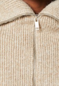 Beiger Strickpullover mit einer gerippten Textur, ausgestattet mit einem silbernen Reißverschluss und einem weichen Kragen. Der Stoff wirkt warm und gemütlich.