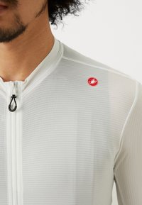 T-shirt de sport léger et blanc avec fermeture éclair et motif rayé texturé. Présente un logo rouge sur l'épaule et une tirette en boucle.
