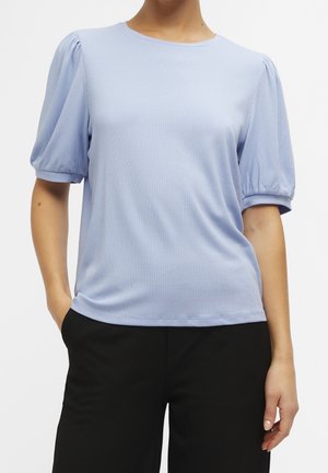 Woman wearing a light blue ribbed knit top with puffed short sleeves and black pants, hands in pockets against a white background.