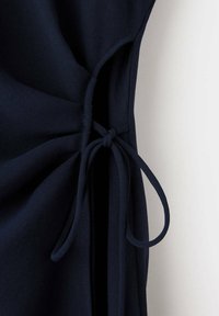 Gros plan d'un tissu bleu foncé noué en nœud avec des bretelles étroites, montrant une fente latérale sur un fond clair.