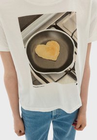 White cotton t-shirt featuring a printed graphic of a heart-shaped food item in a frying pan, set against a kitchen background.