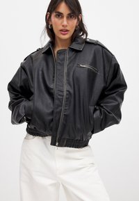 Giacca bomber nera in ecopelle con chiusura zip, orlo a coste e tasca sul petto con zip. Presenta spalline e una vestibilità comoda.