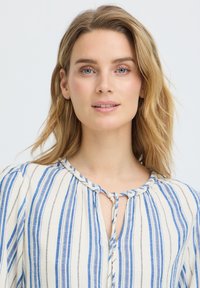 Femme blonde aux yeux bleus portant une blouse blanche à rayures verticales bleues et un col noué, regardant devant elle avec une expression neutre.