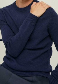 Uomo che indossa un maglione a maglia blu navy con polsini e orlo a coste, con la mano destra appoggiata sulla spalla sinistra.
