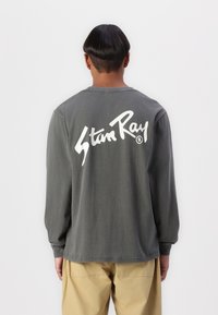 Dlhý rukáv, tričko v tmavošedej bavlne s veľkým bielym logom "Stan Ray" potlačeným na chrbte. Klasický okrúhly výstrih a voľný strih.