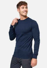 Pull à côtes bleu marine à manches longues, col rond, avec un logo discret au niveau de l'ourlet, associé à un pantalon gris.