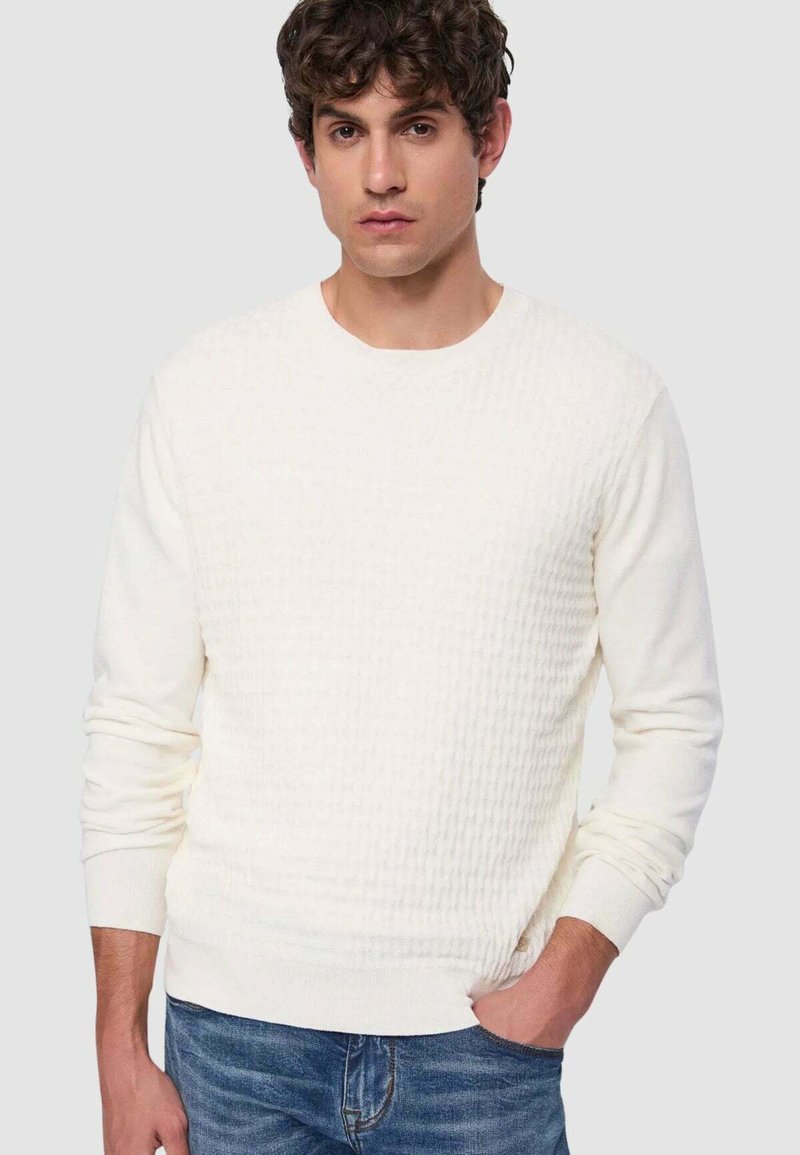 Maglione bianco testurizzato con maniche lunghe e scollo rotondo, abbinato a jeans di denim blu. Tessuto liscio con motivo leggermente in rilievo.