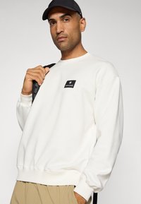 Felpa bianca realizzata in tessuto morbido, con un logo quadrato nero sul petto. Polsini e orlo a costine, abbinata a uno zaino nero.