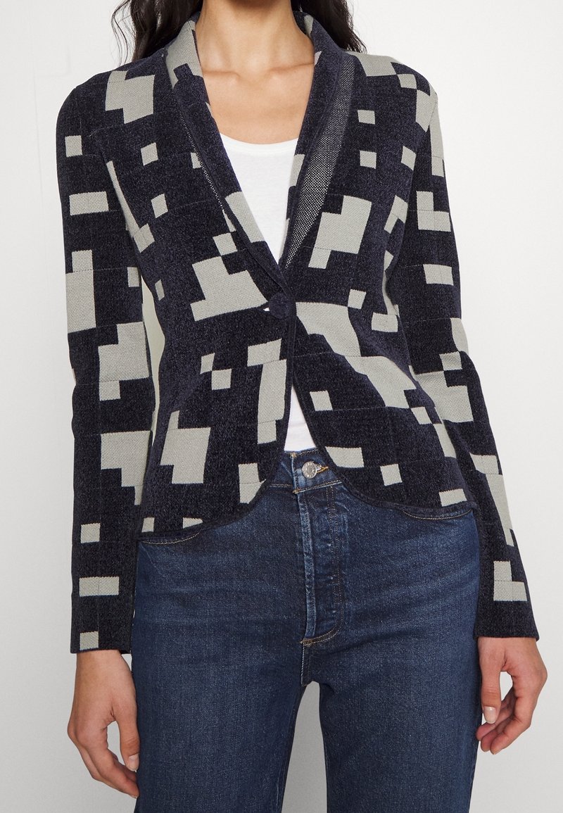 Kvinna klädd i en marinblå och ljusgrå jacka med geometriskt mönster över en vit topp och mörkblå jeans.