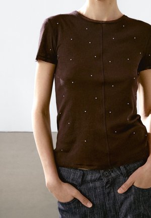 Schlanke Person trägt ein dunkelbraunes kurzärmliges Hemd mit kleinen weißen Punkten und dunkelgraue Hose, die Hände in den Taschen.