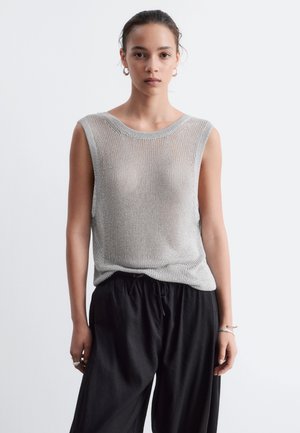 Jeune femme aux cheveux foncés attachés, portant un haut en maille argentée sans manches et un pantalon noir ample, debout devant un fond clair uni.