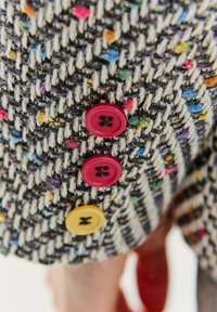 Multicolorová tkanina z tweedu s melírovanou strukturou, doplněná řadou tří knoflíků: dvě červené a jeden žlutý.