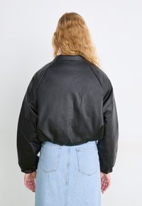 Μαύρο δερμάτινο μπουφάν με χαλαρή εφαρμογή, που διαθέτει λεία υφή και φαρδύ γιακά, συνδυασμένο με μια ανοιχτόχρωμη μπλε τζιν φούστα.