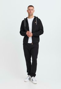 Giovane uomo in piedi con un cappuccio nero con zip, t-shirt bianca, pantaloni neri e scarpe bianche con strisce nere, su uno sfondo semplice.