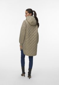 Manteau matelassé beige avec capuche, présentant des manches longues et un ourlet légèrement arrondi. Associé à un jean bleu et des bottes à talons noirs.