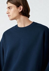 Tamsiai mėlynas megztinis su raižyta apykakle ir nuleistais pečiais. Pagamintas iš minkštos medžiagos, jis turi laisvą siluetą ir glotnią tekstūrą.