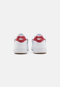 Hvite sneakers med en rød hælaksent, marineblå detaljer og en teksturert overflate. Har en gummisåle og et Lacoste-logo på hælen.