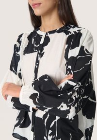 Schwarz-weiße Bluse mit floralen Mustern, puffärmeligen Ärmel und runden Ausschnitt. Der Stoff wirkt leicht und luftig mit Knopfdetails.
