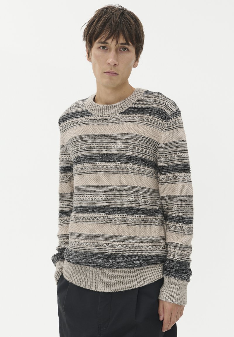 Ung man med brunt hår som bär en beige och svart randig stickad tröja och svarta byxor mot en vit bakgrund.