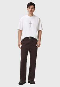 Valkoinen puuvillainen T-paita, jossa on keskeinen graafinen ristisuunnittelu, yhdistettynä ruskeisiin puuvillapantseihin ja mustiin kenkiin. Yksinkertainen, rento istuvuus.