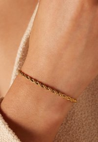 Gouden gedraaid armband om een pols, met een gestructureerd ontwerp van verstrengelde schakels. Gladde afwerking met een subtiele glans.