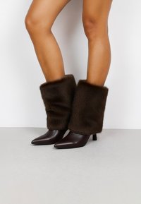  calentadores de piernas de pelaje sintético marrón combinados con botas de tacón oscuro y puntiagudas, que presentan una textura suave y un diseño ajustado.