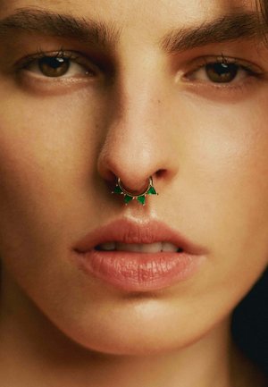 Aro de nariz de oro con tres acentos triangulares verdes. Piel suave y labios naturales, destacando los detalles de la joyería.