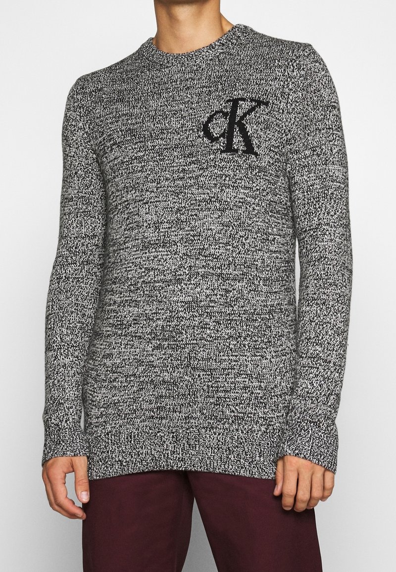Homme portant un pull en maille marbrée noir et blanc avec le logo « CK » sur la poitrine et un pantalon bordeaux foncé, debout sur un fond blanc.